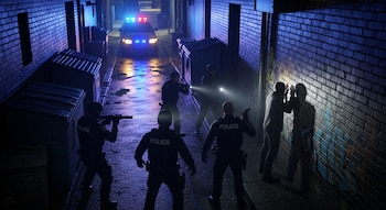 Cinco agentes de policía detienen a un hombre contra una pared en un callejón oscuro y mojado, iluminado por luces de patrulla y linternas.