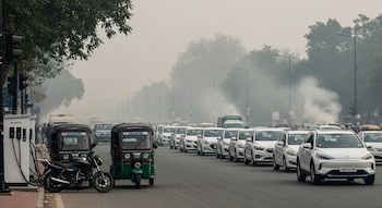 Una avenida con tráfico de vehículos, triciclos y motocicletas. Se observa una estación de carga, árboles y postes de luz bajo un cielo cubierto por smog.