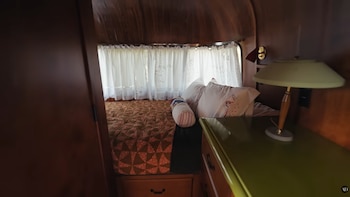 Vista de un dormitorio compacto con cama, colcha estampada, almohadas, cortinas blancas y una lámpara de mesa sobre un mostrador verde