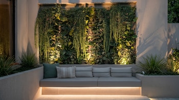 Sofá integrado de color claro con cojines frente a un jardín vertical exuberante y plantas en macetas, iluminado por luces cálidas en un patio moderno.