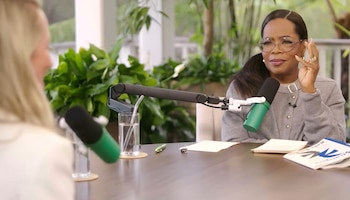 Oprah Winfrey sentada en una mesa, con un micrófono verde y negro enfrente, gesticulando con la mano derecha. Viste un suéter gris y gafas