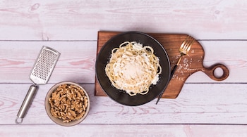 Un cuenco negro con espagueti y salsa, una tabla de madera, un tenedor dorado, un rallador de metal, un cuenco con nueces, una mesa de madera.