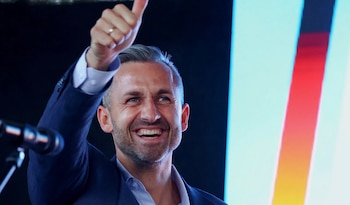 Un hombre con traje sonríe y levanta el pulgar junto a un micrófono y una pantalla con franjas de la bandera de Alemania