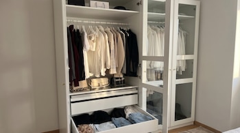 Un armario blanco muestra ropa colgada en perchas, bolsos sobre una bandeja de cristal, un cajón inferior abierto con prendas dobladas y botas visibles detrás de puertas de cristal.