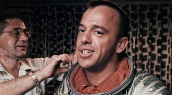 Primer plano de Alan Shepard sonriendo, con un técnico ajustándole el cuello de su traje espacial plateado con forro naranja