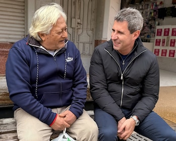 Dos hombres sentados en una banca. Un hombre de cabello blanco viste un buzo azul. Sergio Uñac, de cabello gris, viste una chaqueta negra acolchada