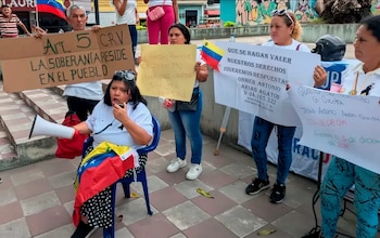 Varias personas en una plaza sostienen carteles y banderas de Venezuela, una mujer sentada habla por megáfono. Los carteles contienen texto en español