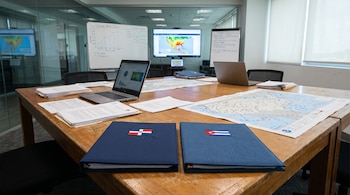 Mesa de madera con documentos, laptops, mapas meteorológicos y carpetas con banderas de República Dominicana y Cuba. Al fondo, pantallas con mapas climáticos.