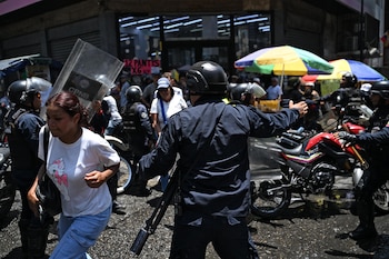 Agentes de policía con cascos y uniformes oscuros en motocicletas en una calle, una mujer de camiseta blanca y jeans camina en primer plano. Hay sombrillas de colores
