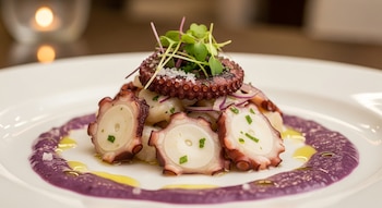 Plato blanco con rodajas de pulpo, cebolla morada, brotes verdes, sal, cama de ingredientes blancos, salsa morada y gotas de aceite.