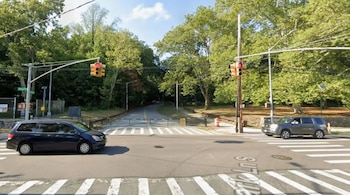Vista de una intersección con semáforos en rojo, dos coches detenidos, un paso de cebra y un camino bloqueado por barreras, con árboles verdes al fondo