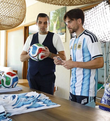 Dos hombres interactúan. Uno con camiseta argentina, marcador en mano; el otro con chaleco, balón de fútbol. Más balones y camisetas dobladas en una mesa