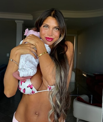 Mujer de cabello largo en traje de baño sostiene en brazos a un bebé vestido de blanco con detalles rosados