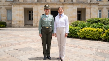 Dos mujeres posan de pie en un patio pavimentado frente a un edificio de piedra con columnas; una viste uniforme militar y la otra ropa civil.