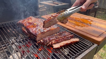 Carne asada rebanada sobre una parrilla con brasas incandescentes, una mano que sostiene un trozo con pinzas y una tabla de madera.