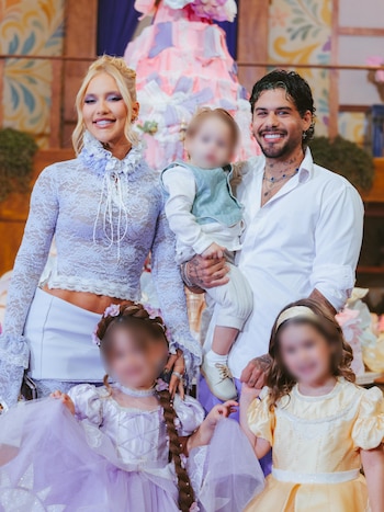 Una mujer rubia con blusa de encaje lila, un hombre con camisa blanca que carga a un bebé y dos niñas con vestidos ante un fondo decorado