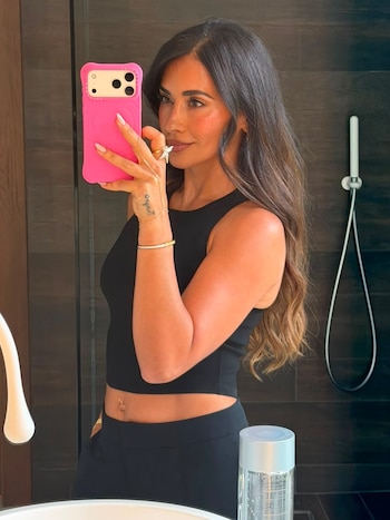 Antonela Roccuzzo sostiene un teléfono con funda rosa frente a un espejo y viste un top corto sin mangas de color negro