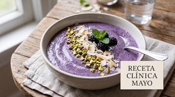 Un tazón con licuado morado decorado con moras frescas, pistaches picados y coco sobre una mesa de madera.
