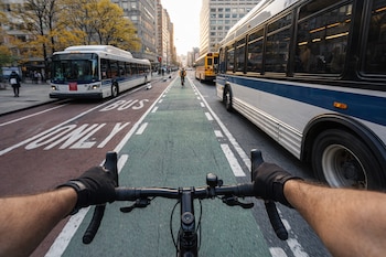 Vista en primera persona de un ciclista pedaleando por un carril verde en Nueva York, flanqueado por dos autobuses blancos y azules.