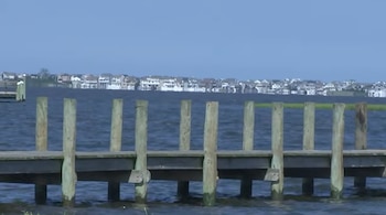 Muelle de madera sobre cuerpo de agua, con postes verticales. Al fondo, una hilera de casas sobre la orilla opuesta bajo cielo azul claro