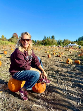 Una mujer con gafas de sol y sudadera posa sentada sobre una gran calabaza en un campo agrícola lleno de calabazas dispersas