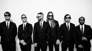 Seis miembros de Maroon 5 de pie en blanco y negro, vestidos con trajes negros, camisas blancas, corbatas negras y gafas de sol oscuras, contra un fondo blanco