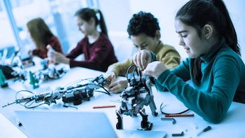 Varios niños sentados a una mesa blanca ensamblan dispositivos robóticos con piezas de plástico y cables en un aula
