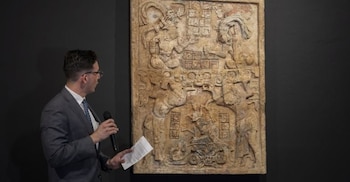 Un hombre con traje y gafas habla en un micrófono junto a un gran relieve de piedra antiguo con intrincadas figuras y jeroglíficos en una pared oscura