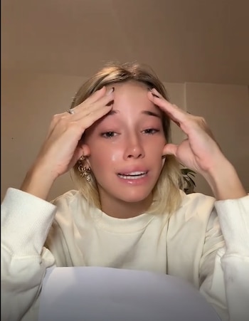 Zoe Bogach, una joven rubia, llorando con lágrimas visibles en sus mejillas, sus manos en las sienes y expresión de angustia, con fondo claro