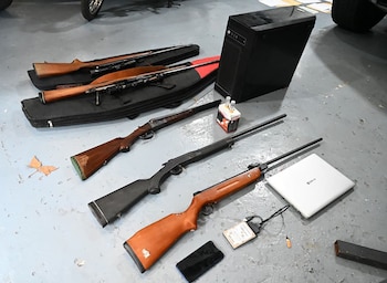 Surtido de armas de fuego largas, una computadora, un portátil, un smartphone y un objeto blanco pequeño con tubos sobre un suelo gris