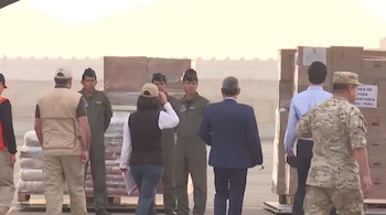Grupo de personas en uniforme militar y ropa de civil junto a cajas de cartón apiladas en paletas sobre el asfalto en una pista de carga