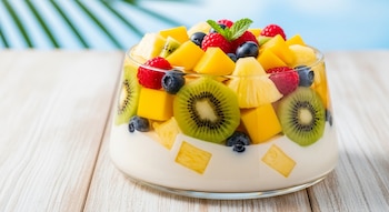 Un vaso de vidrio con un postre de gelatina de coco cubierto con trozos de mango, piña, kiwi, frambuesas y arándanos, sobre una mesa de madera clara.