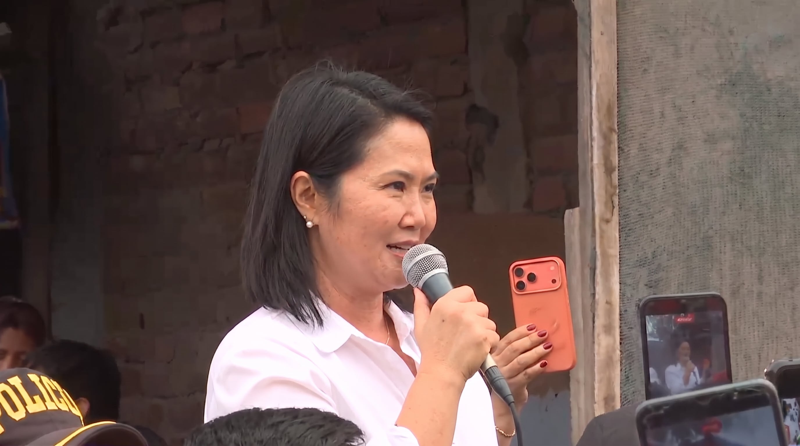 Keiko Fujimori habla ante una audiencia, sosteniendo un micrófono y un teléfono móvil mientras otros la graban con sus dispositivos. (Imagen Ilustrativa Infobae)