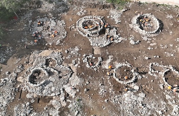 Vista aérea de personas con chalecos naranjas que trabajan entre estructuras circulares de piedra y tierra divididas en cuadrículas