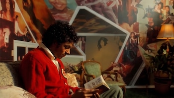 Michael Jackson joven, con cárdigan rojo y camisa blanca, sentado en un sofá leyendo un libro. Al fondo, una pared decorada con fotografías y cuadros