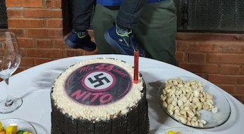 Una torta de chocolate con esvástica, texto Mis 44 años Nito y una vela roja; una copa vacía, un plato con bocadillos, y piernas con zapatillas
