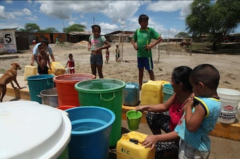Varias personas y niños llenan baldes y bidones plásticos de agua en un terreno de tierra junto a viviendas y un perro