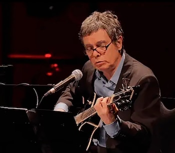 Un hombre de mediana edad con cabello gris y gafas toca una mandolina negra y canta en un micrófono sobre un escenario oscuro