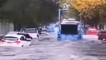 Imagen borrosa de una calle inundada con varios automóviles sumergidos casi hasta las ventanillas y un vehículo más grande en el centro que desplaza agua