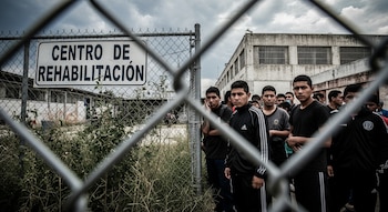 Grupo de jóvenes visto a través de una cerca metálica, junto a un letrero que dice 'CENTRO DE REHABILITACIÓN'. Edificios con ventanas enrejadas bajo un cielo nublado.
