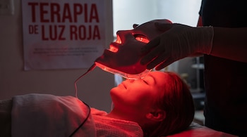 Manos con guantes sostienen una máscara que emite luz roja sobre el rostro de una persona acostada junto a un cartel de terapia de luz roja.