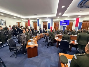 Vista amplia de una sala de conferencias con decenas de personas, muchas en uniformes militares o policiales, de pie frente a una mesa curva. Una pantalla muestra un texto de certificación