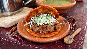 Gordita de maíz con carne deshebrada, col picada, cebolla, cilantro y queso fresco sobre un plato de barro con chiles secos alrededor.