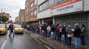 Larga fila de personas usando mascarillas frente a la Oficina de Empleo SENA en Bogotá, con taxis amarillos en la calle mojada.