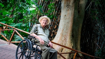 Hombre mayor en silla de ruedas, con sombrero y chaleco, en un entorno natural con abundante vegetación. A su derecha, un árbol con las iniciales 'JEBE 1964' grabadas