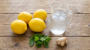 Cuatro limones, una jarra de vidrio con agua e hielo, hojas de menta y una raíz de jengibre sobre una mesa de madera.