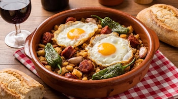 Una cazuela de barro con migas de pan, dos huevos fritos, rodajas de chorizo, pimientos verdes y ajos, junto a un trozo de pan y vino tinto.