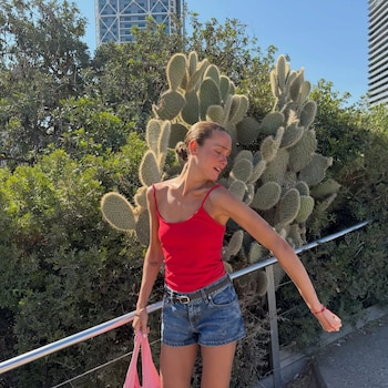 Una mujer joven con cabello recogido viste top rojo y short de jean, sostiene una bolsa rosa, posa junto a un cactus, un arbusto y un edificio moderno