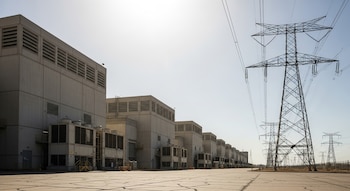 Una fila de edificios de centros de datos bajo el sol intenso. Pavimento agrietado en primer plano y varias torres eléctricas al fondo.