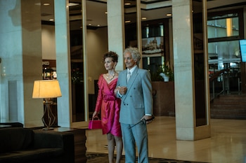 Un hombre con traje gris y radiotransmisor camina tomado del brazo de una mujer con vestido fucsia en el vestíbulo de un hotel